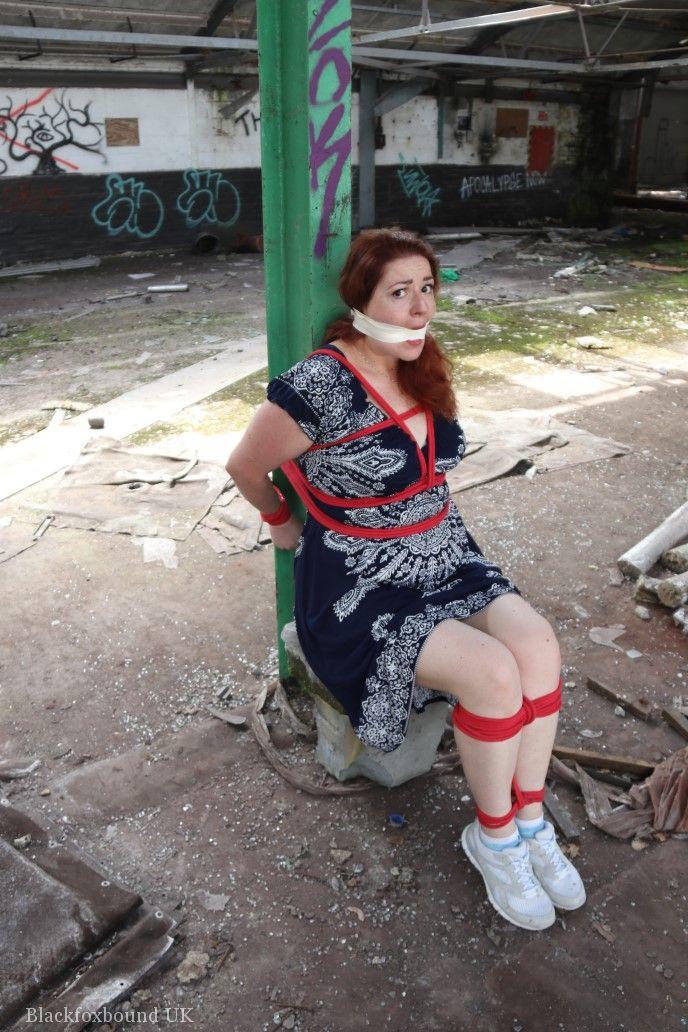 倉庫で服を着たまま口を塞がれ柱に縛られた赤毛のぽっちゃり女性