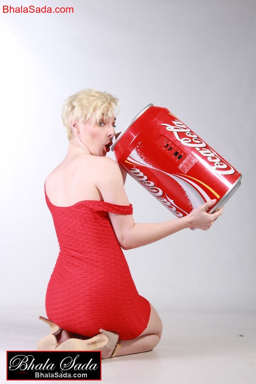 أمّ ناضجة ذات شعر قصير في فستان أحمر جذاب تشرب الكوكاكولا من علبة ضخمة